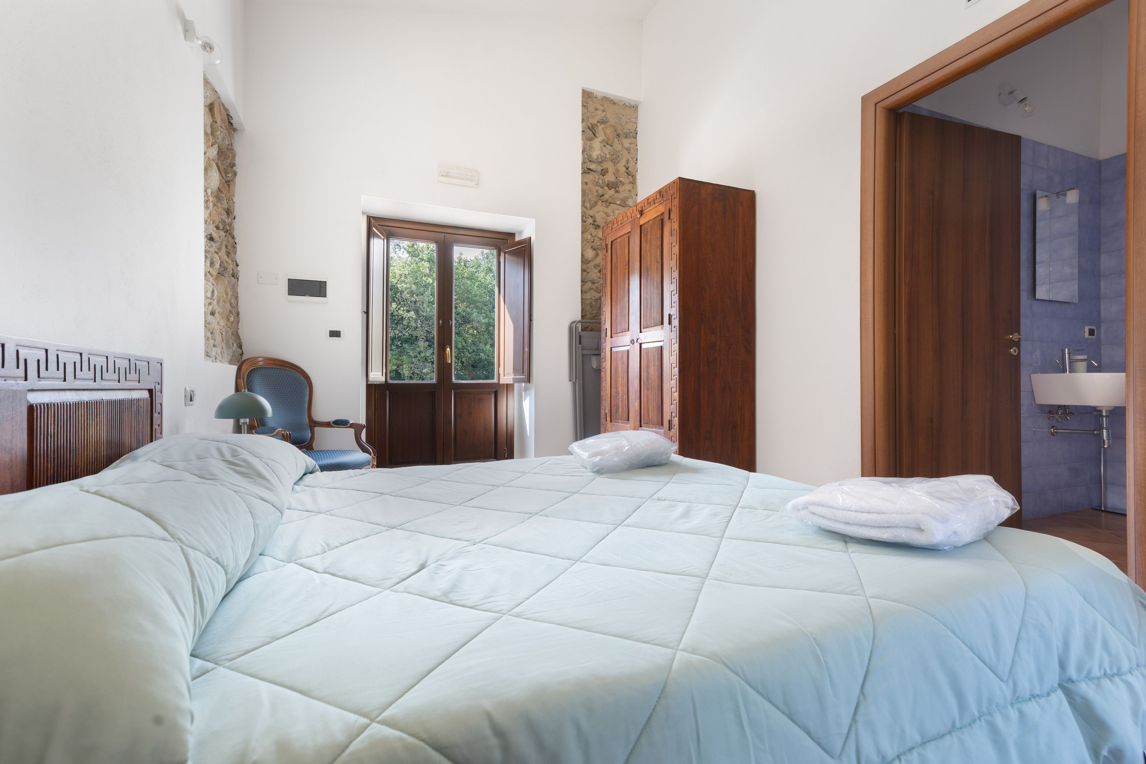 Une chambre à coucher avec un grand lit, une chaise, une armoire en bois et une salle de bain visible à travers une porte ouverte.