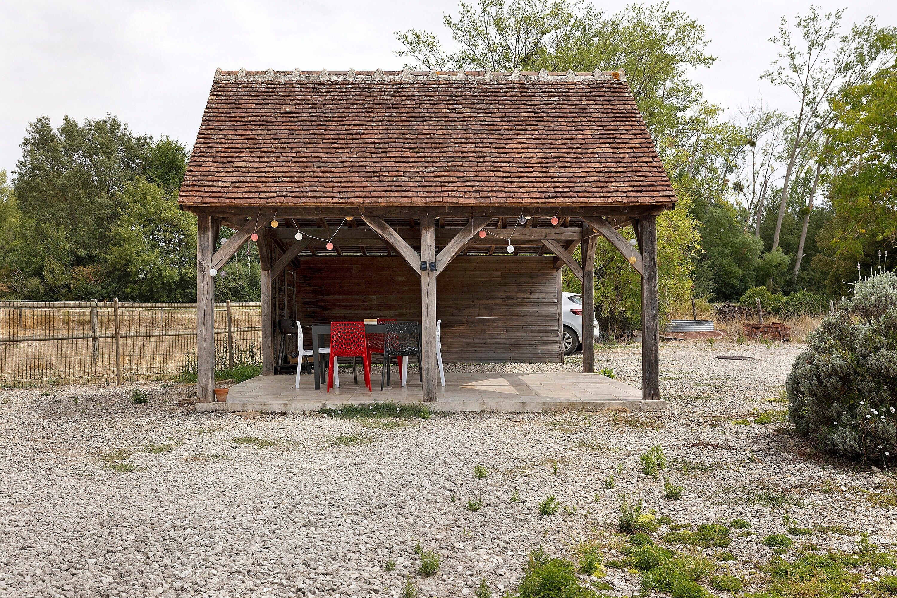 A rustic wooden pavilion with a tiled roof, surrounded by gravel, featuring a seating area with chairs and a table.