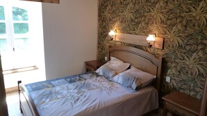 Un dormitorio con una cama, dos mesitas de noche, una mesilla auxiliar, un ventanal y una pared con estampado floral.