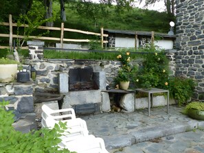 A stone-built outdoor seating area with a stone fireplace, surrounded by greenery and a wooden fence.