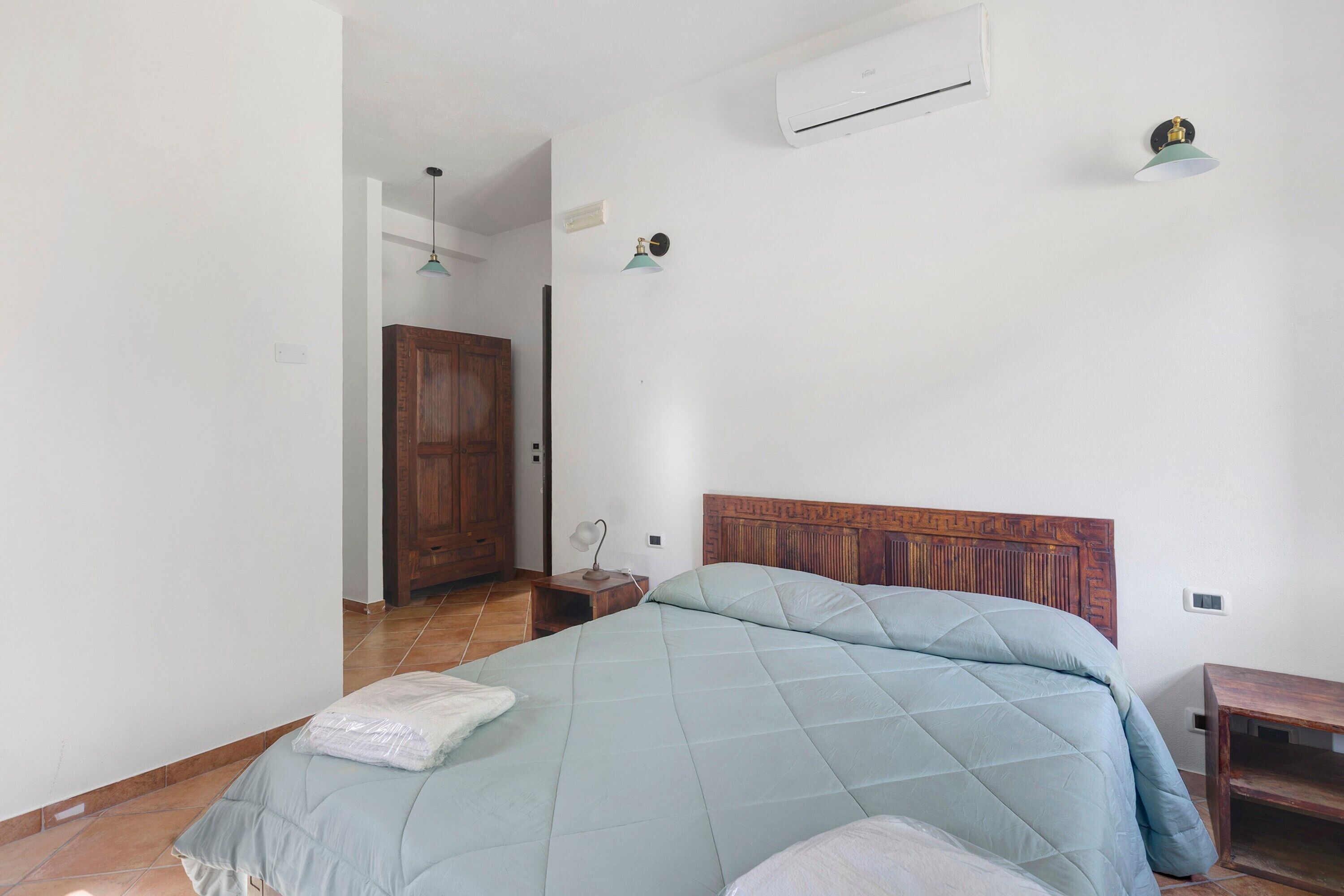 A bedroom with a bed, a wooden wardrobe, an air conditioning unit, and a nightstand with a lamp.
