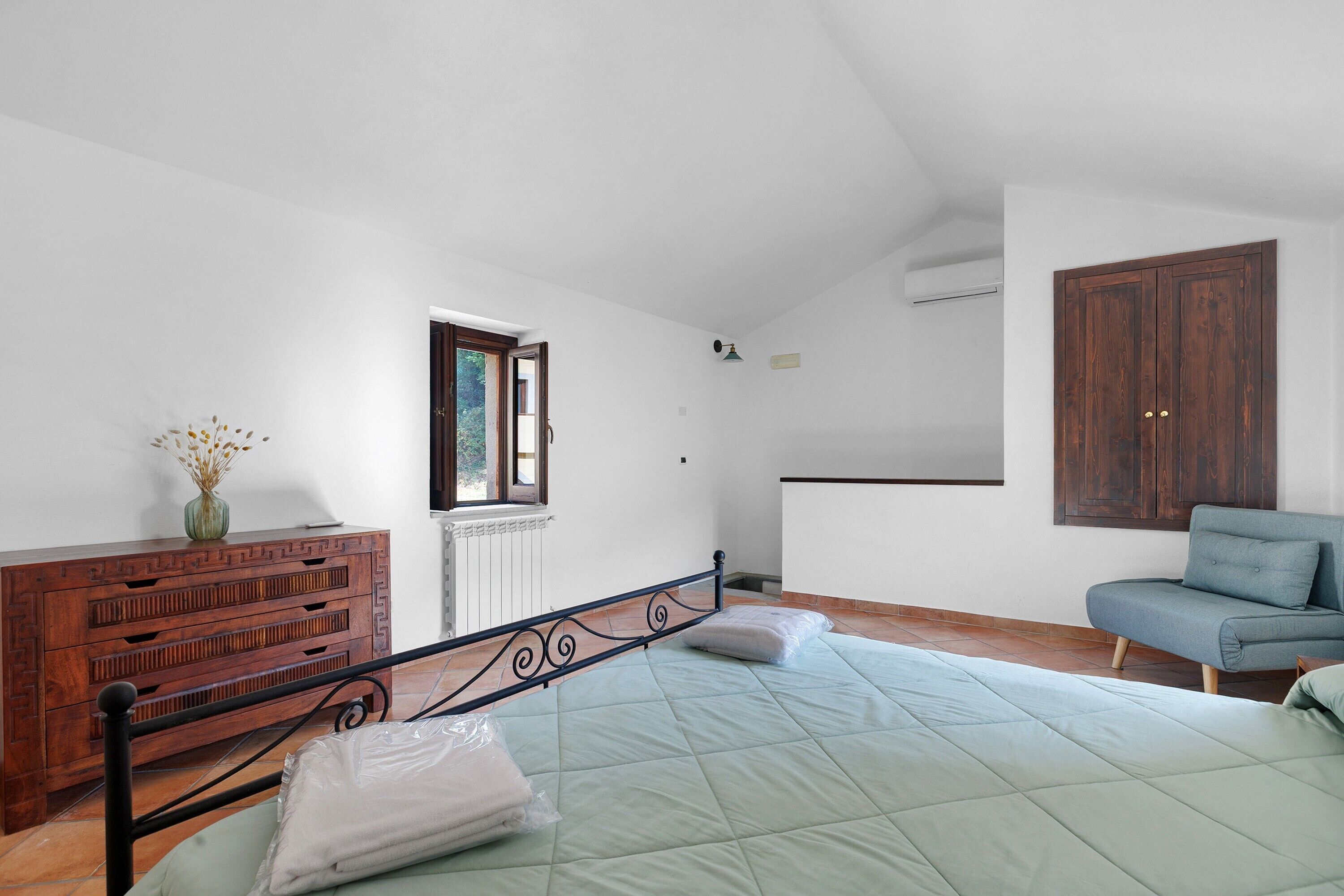 Une chambre à coucher avec un lit, une commode en bois, une fenêtre, un radiateur, un climatiseur mural et un canapé bleu.