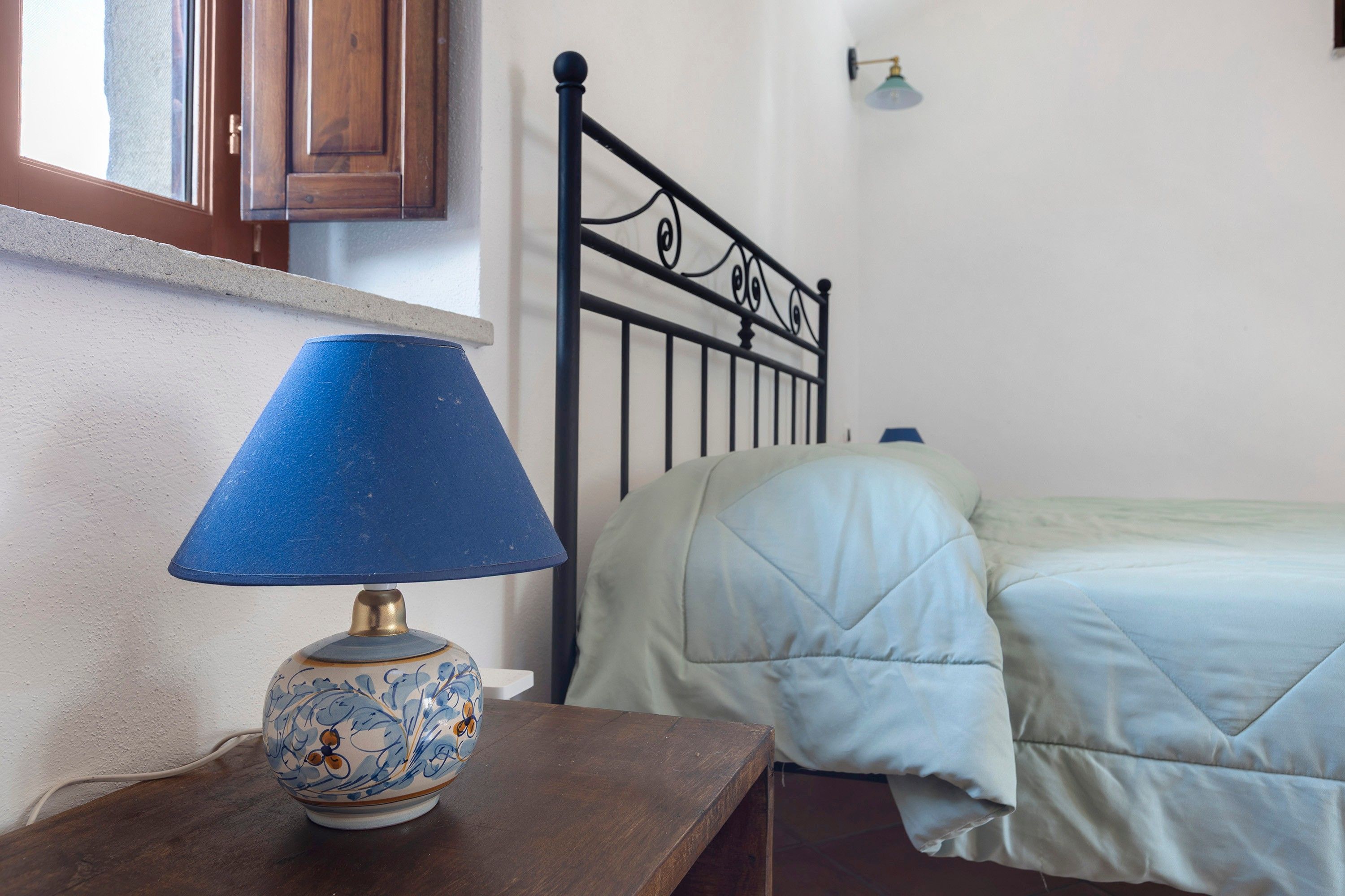 Quarto com mesa de cabeceira de madeira, abajur azul, cama com colcha branca e cabeceira de metal preto.