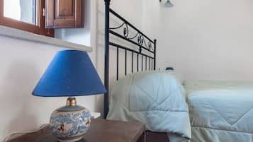 Quarto com mesa de cabeceira de madeira, abajur azul, cama com colcha branca e cabeceira de metal preto.