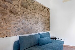 Habitació amb paret de pedra, llit amb roba de llit blava i taula petita.