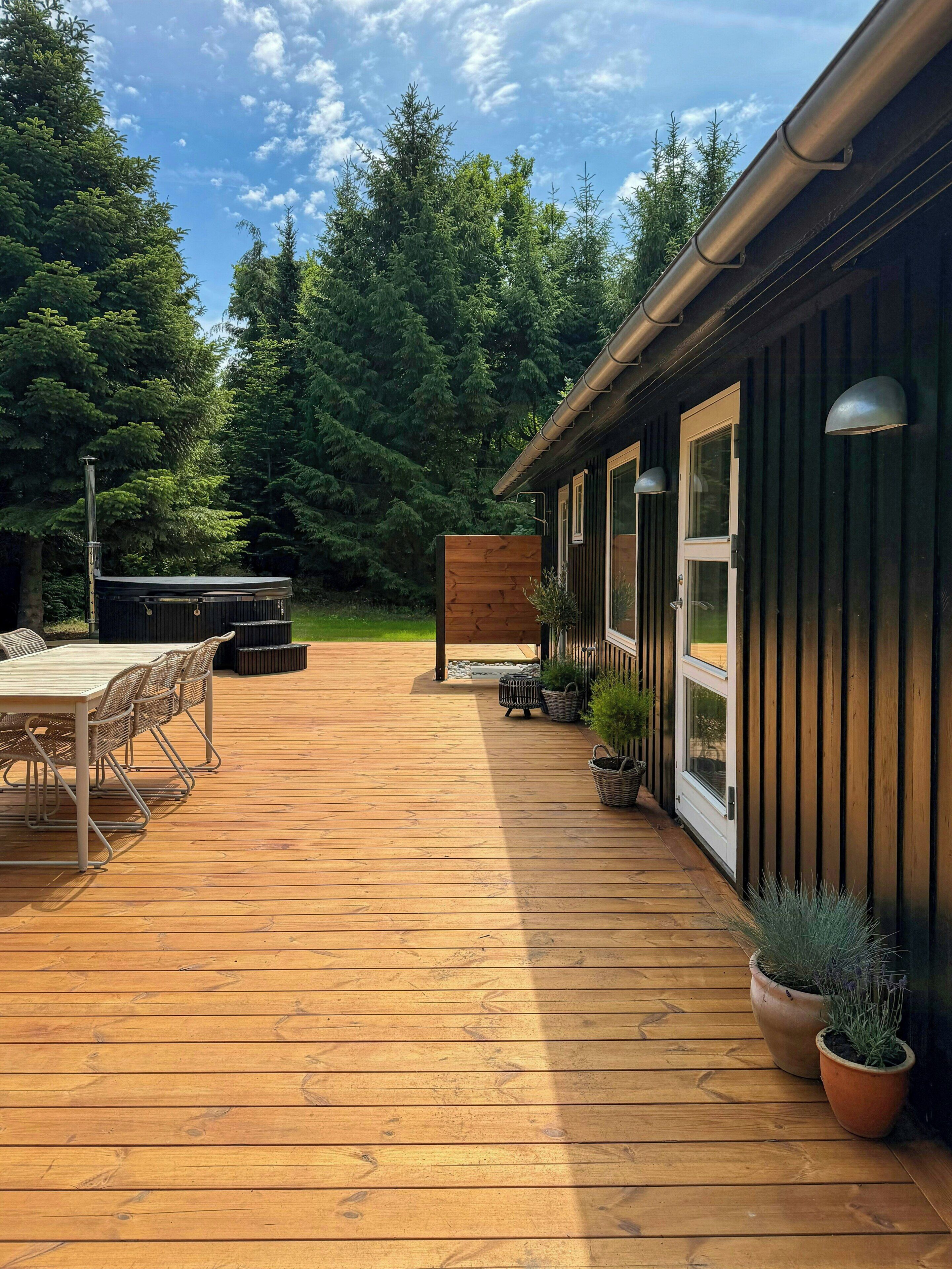 En træterrasse med et bord og stole, omkranset af plantekasser, der fører hen til en bygning med store vinduer og en dør.