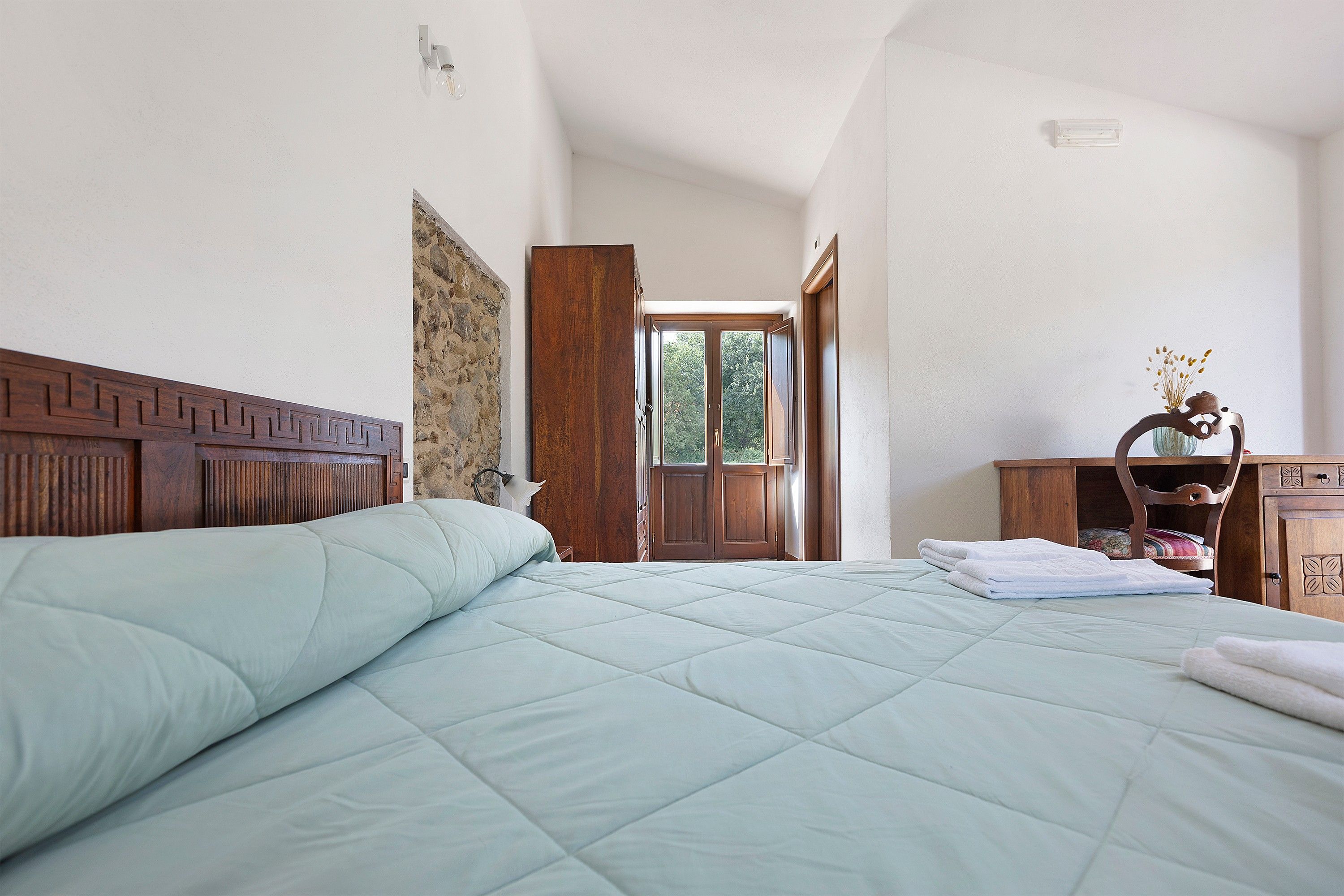 Une chambre à coucher avec un lit, une tête de lit en bois, une table de chevet et une vue sur l’extérieur, visible à travers une porte ouverte.