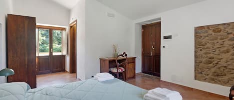 A bedroom with a bed, a wooden wardrobe, a desk with a chair, and a stone wall.