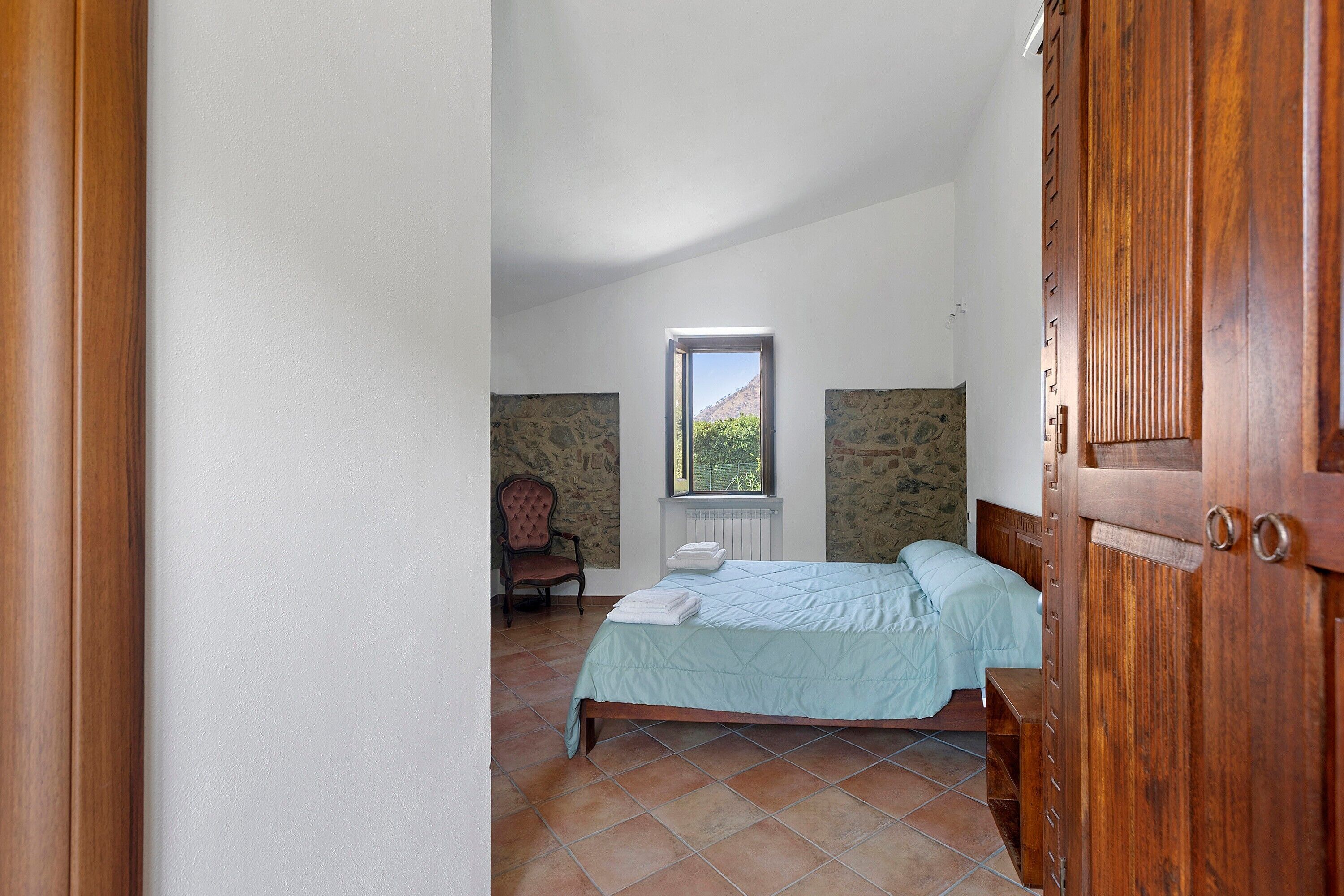 A bedroom with a bed, a chair, a window, and a view of the outdoors.