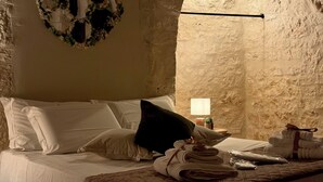 Ein Bett mit weißen Leinen, einem dunklen Kopfkissen und einer gesteppten Decke. Ein kleiner Tisch mit einer Lampe und einem Topfpflanzen.