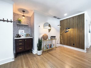 Un interior moderno con suelo de madera, un mueble de madera oscura, una pared blanca con papel tapiz estampado, una estantería con objetos decorativos, un espejo y un radiador fijado a la pared.
