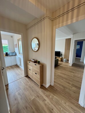Ein geräumiges Zimmer mit Holzboden, einer Holzkommode, einem runden Spiegel und Blick in einen anderen Raum mit Fernseher und Pflanze.