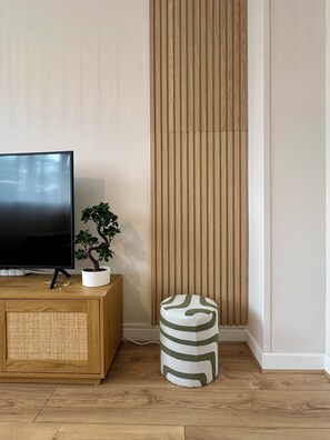 A flat-screen TV on a wooden cabinet, a potted plant, a vertical wooden panel, and a striped ottoman on a wooden floor. - L'Escale Nippone – Wi-Fi – Free Parking (Vichy)