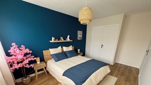 A bedroom with a bed, bedside tables, a nightstand, a hanging light fixture, a wall-mounted shelf with decorative items, and a door. - L'Escale Nippone – Wi-Fi – Free Parking (Vichy)