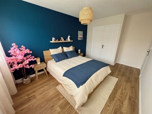 Ein Schlafzimmer mit Bett, Nachttischen, einer Wandlampe, einem an der Wand befestigten Regal mit Dekoration und einer Tür.