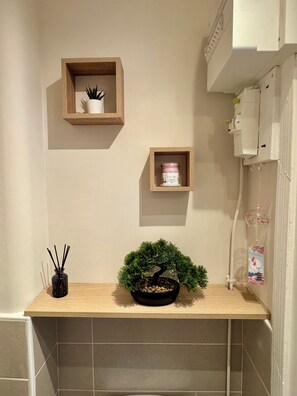 A bathroom with a wooden shelf holding a potted plant, a small white container, and a black bottle with a reed diffuser. - L'Escale Nippone – Wi-Fi – Free Parking (Vichy)