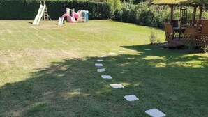 Een gazon met een speeltoestel, een houten terras en een rieten kiosk.
