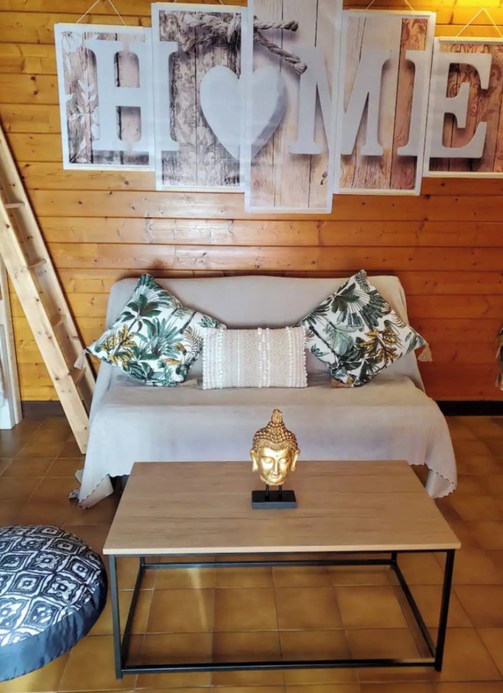 A cozy room with a wooden wall, a sofa with patterned cushions, a small table with a Buddha statue, and a 'HOME' sign above.