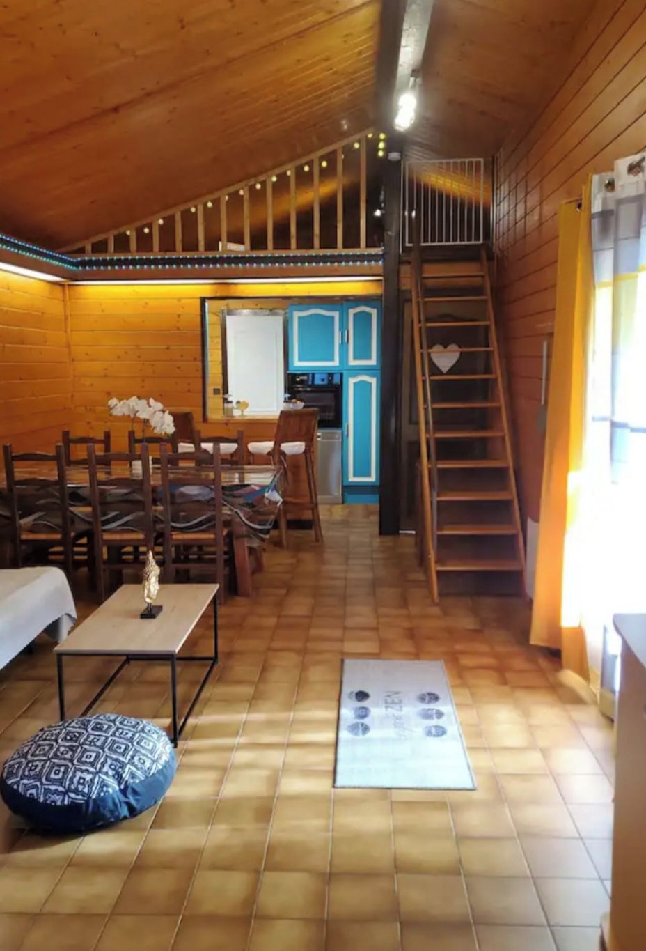 Un accogliente interno di una baita con zona pranzo, angolo cottura e soppalco con scala in legno.