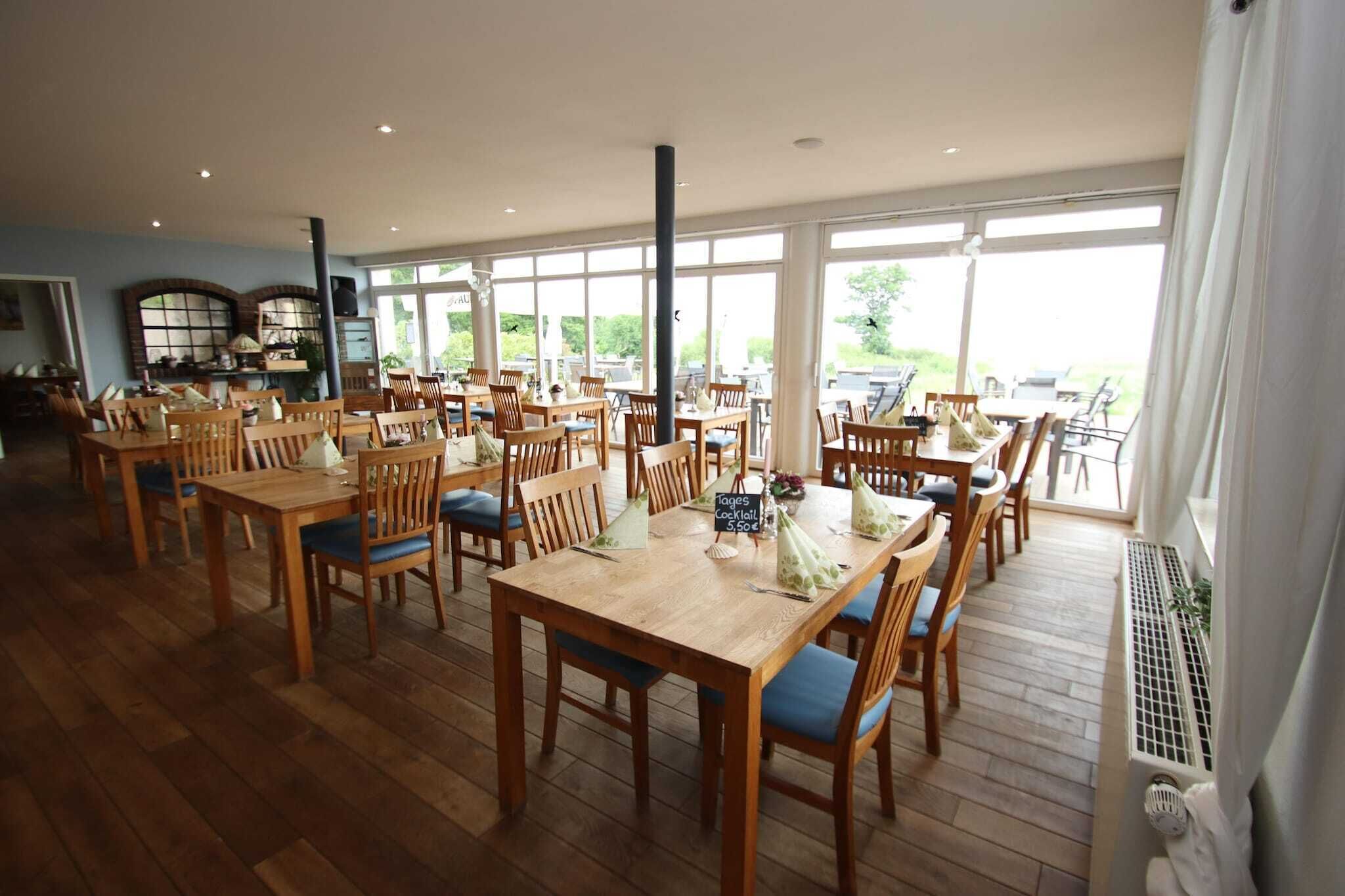 A spacious dining area with multiple wooden tables and chairs, large windows offering a view of the outdoors, and a central column.