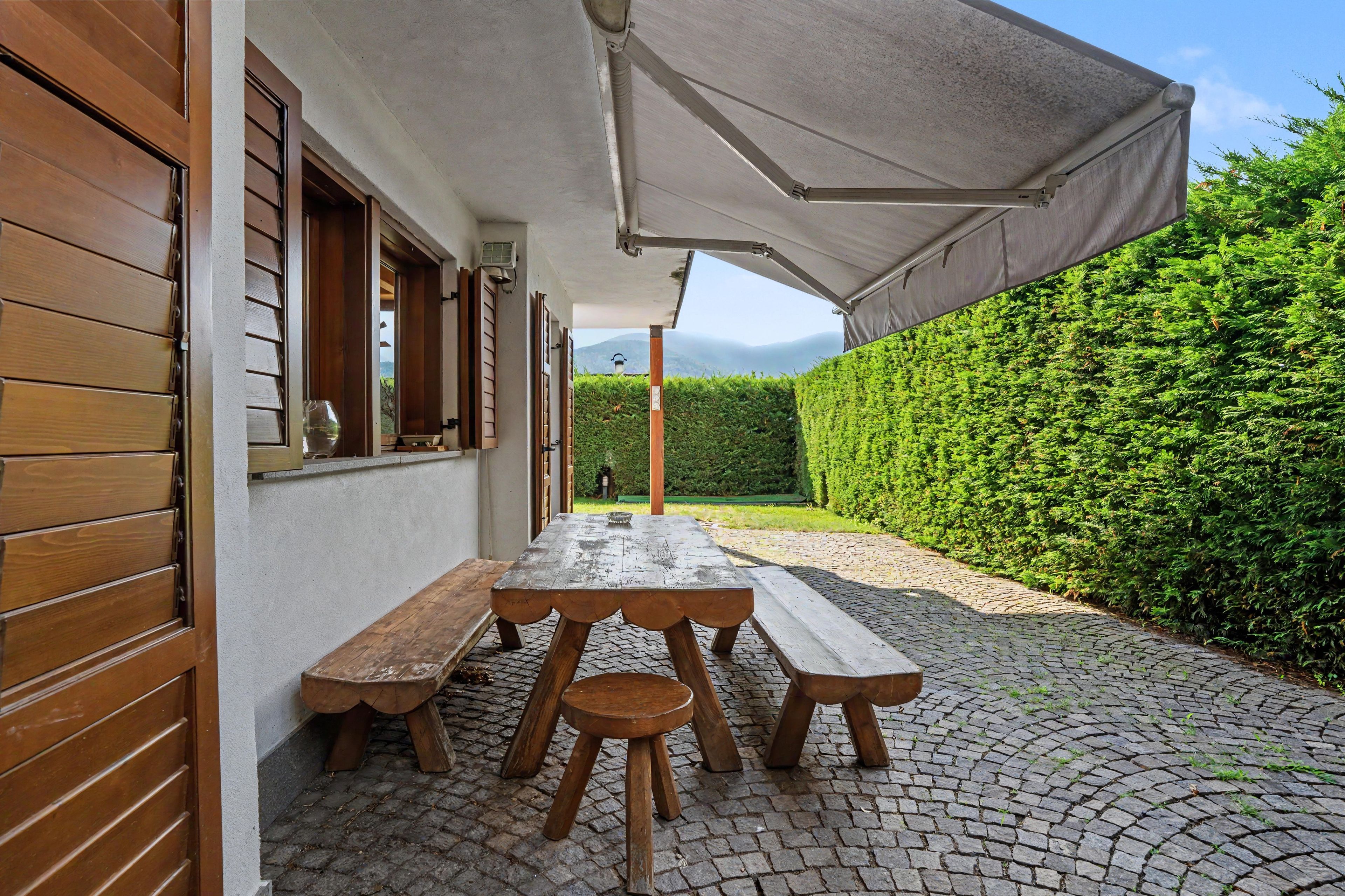 Eine Terrasse mit einem Holztisch und Bänken, einem überdachten Bereich und einer Hecke im Hintergrund.