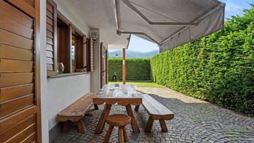 A patio with a wooden table and benches, a covered area, and a hedge in the background.