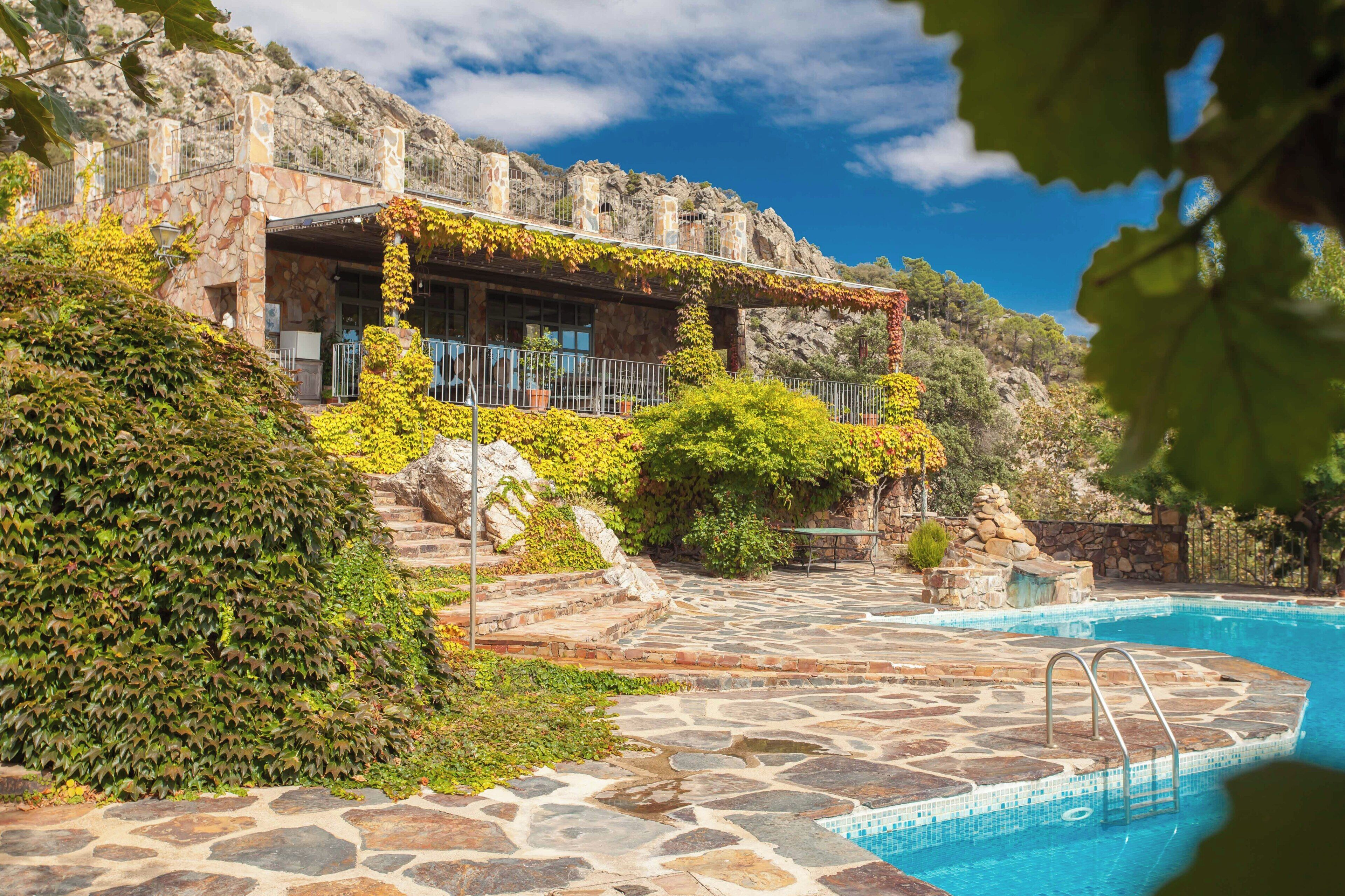 A stone pool with a stone deck leading to a building with a stone facade and a covered patio area.