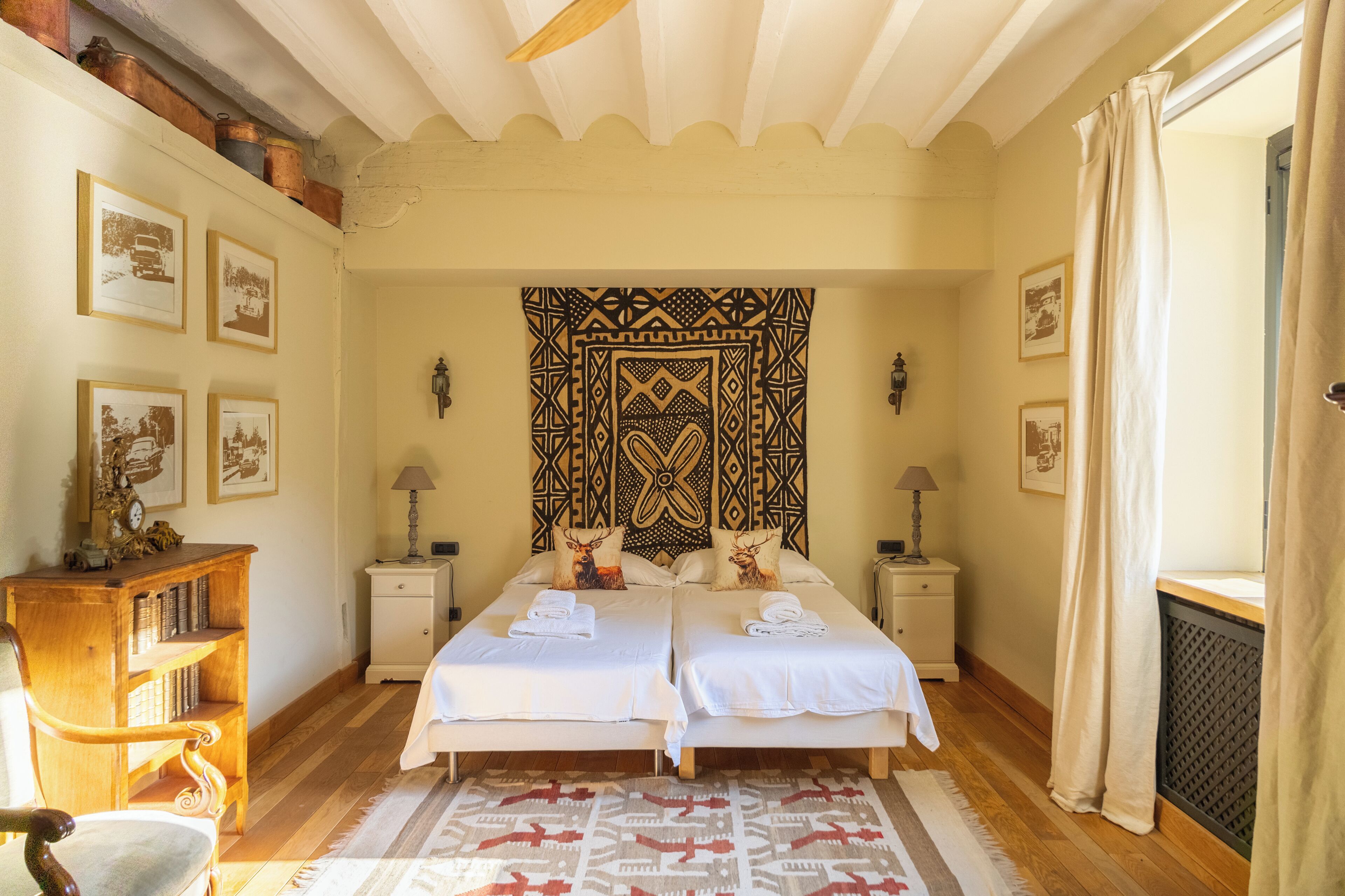 Een tweepersoonsbed met twee kussens, een geborduurd wandkleed, bedlampjes en een houten nachtkastje met boeken.