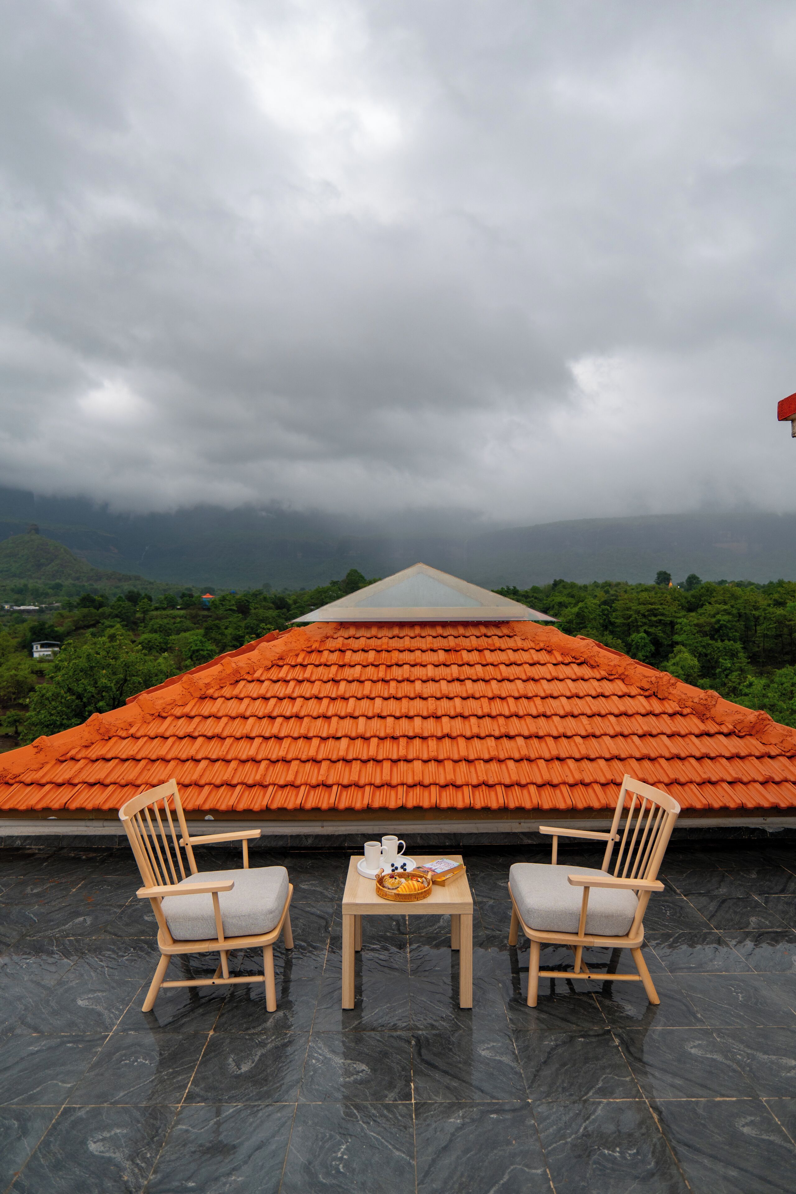 Une terrasse avec deux chaises en bois et une table, un toit en tuiles et une vue sur des collines verdoyantes sous un ciel nuageux.