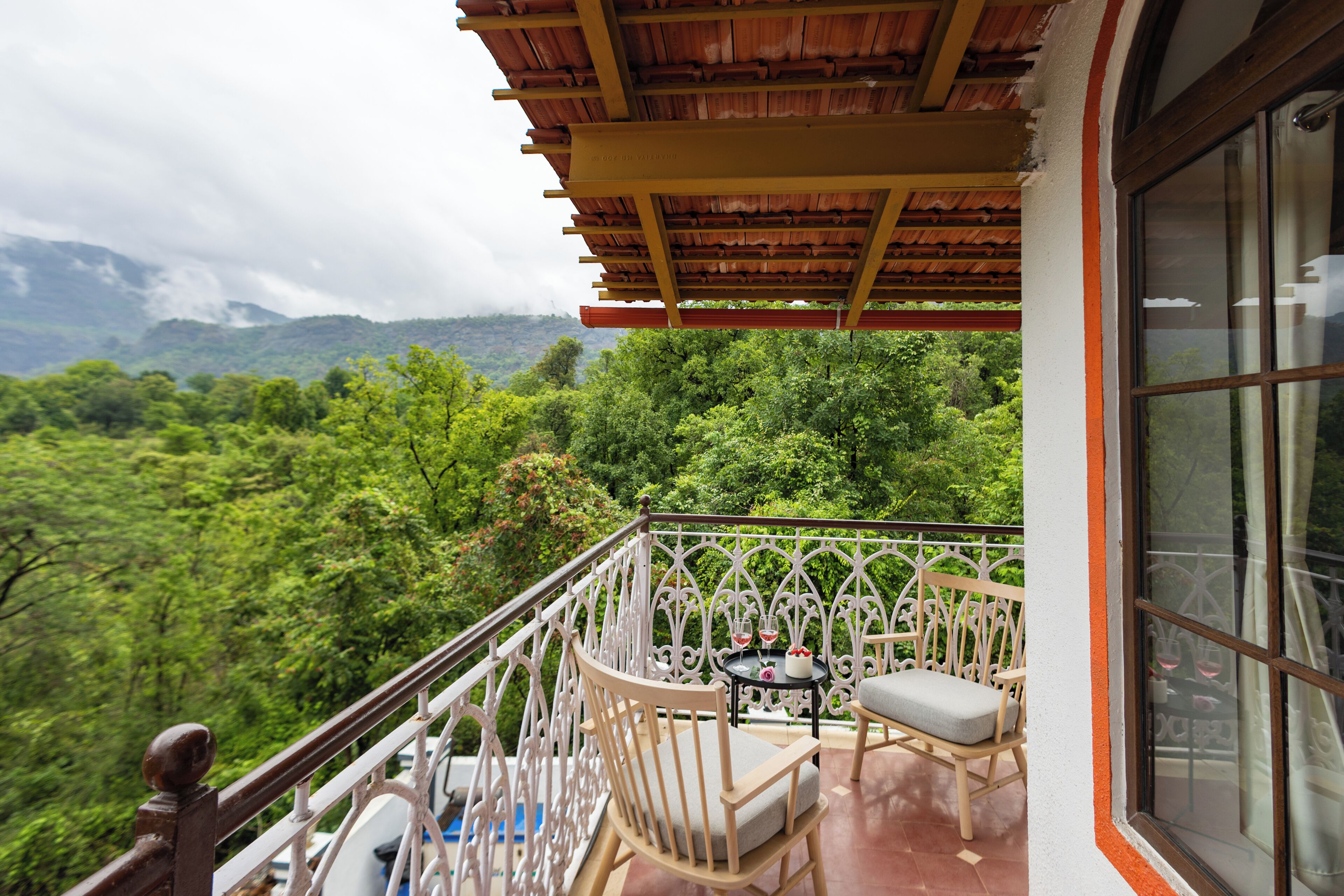 Un balcón con vistas a un paisaje verde exuberante, una mesa con sillas y una ventana con cortinas.
