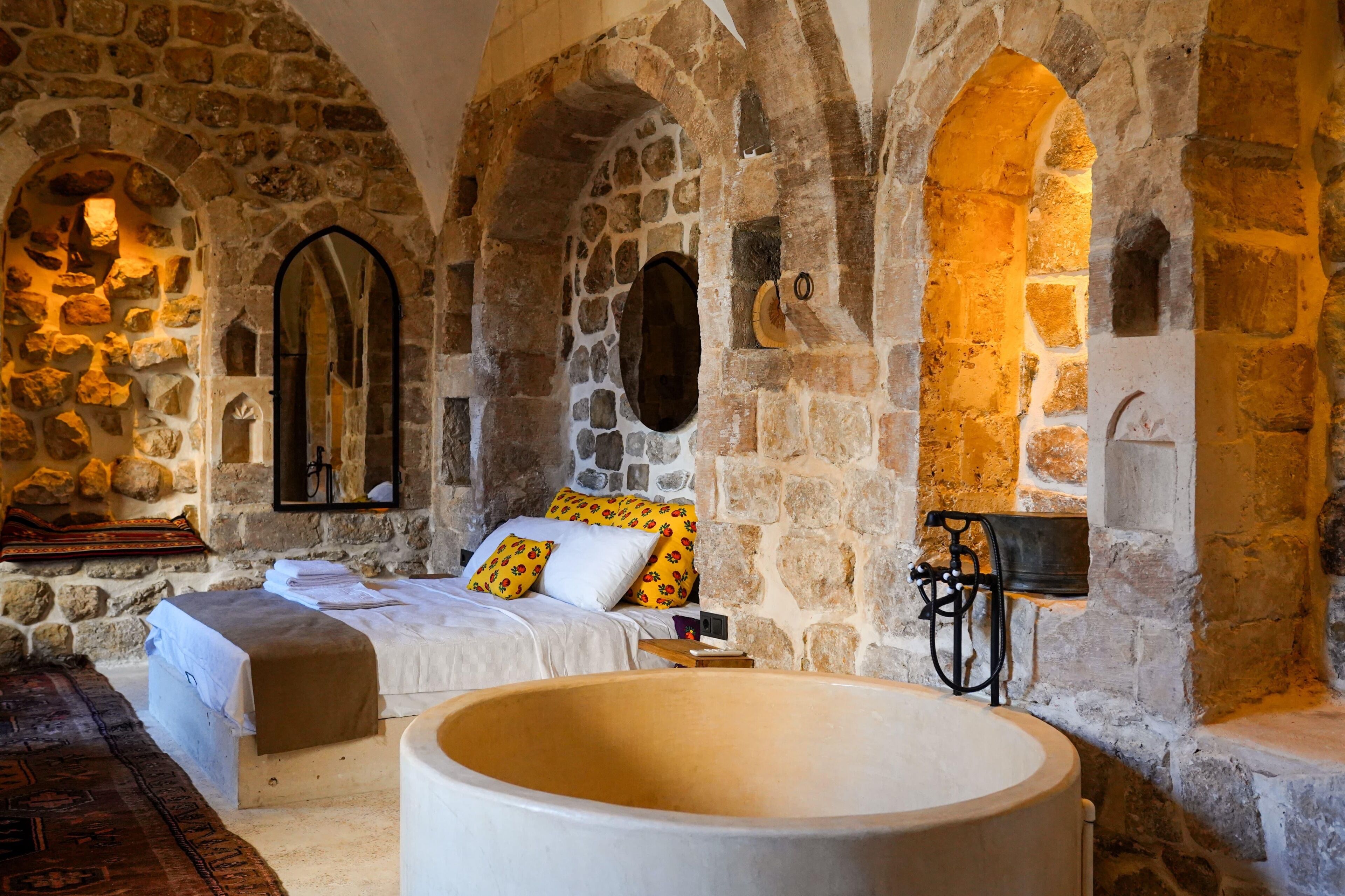 Habitación de paredes de piedra con cama, una bañera grande y un banco de madera.