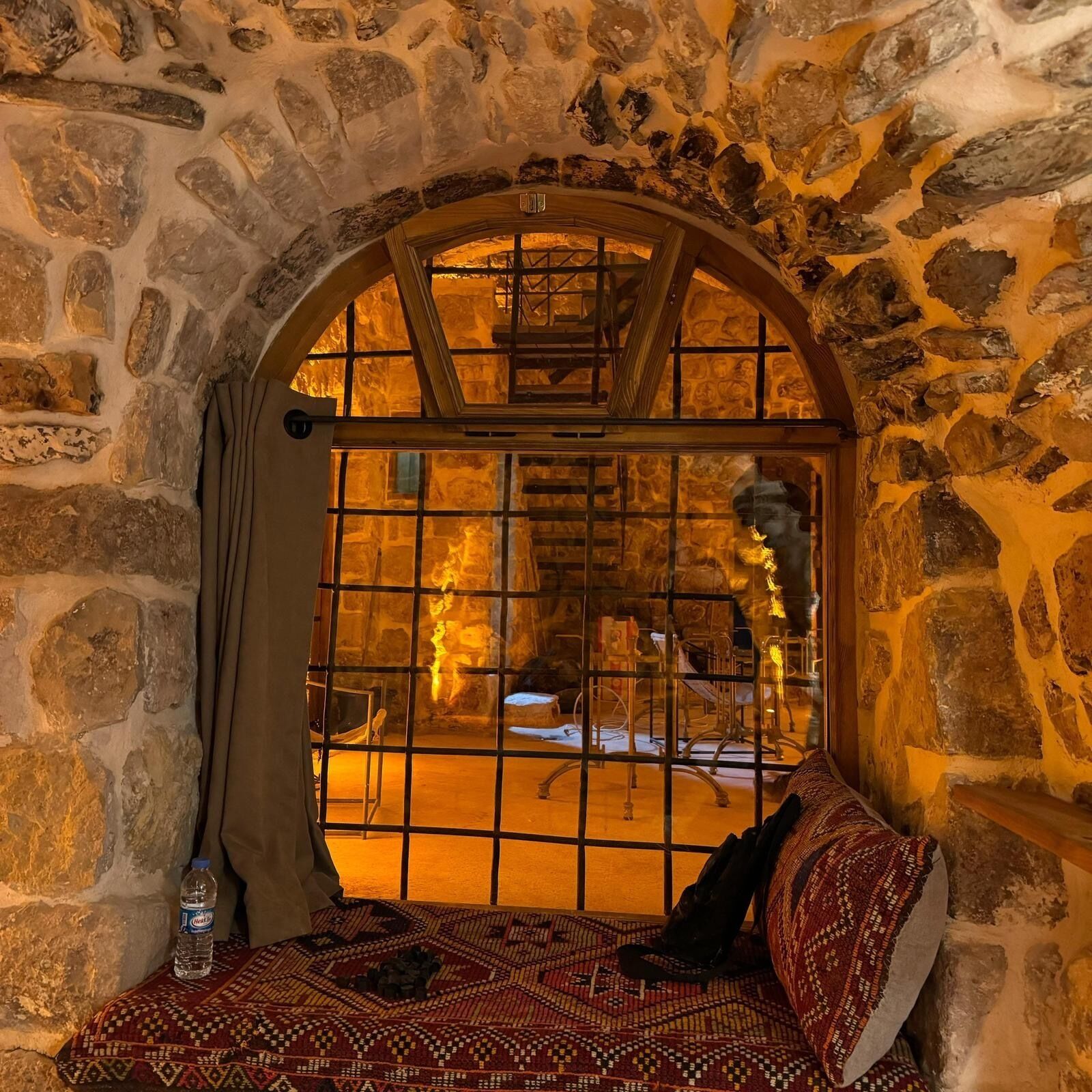 Una habitación de paredes de piedra con un banco, una ventana con barrotes y una botella sobre el banco.