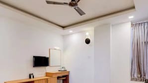A hotel room with a bed, a desk, a television, and a ceiling fan. - NA Grand (Chennai)