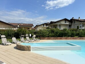 Ein Swimmingpool mit Liegestühlen und einer Steinsäule, umgeben von Grünflächen und Wohnhäusern.