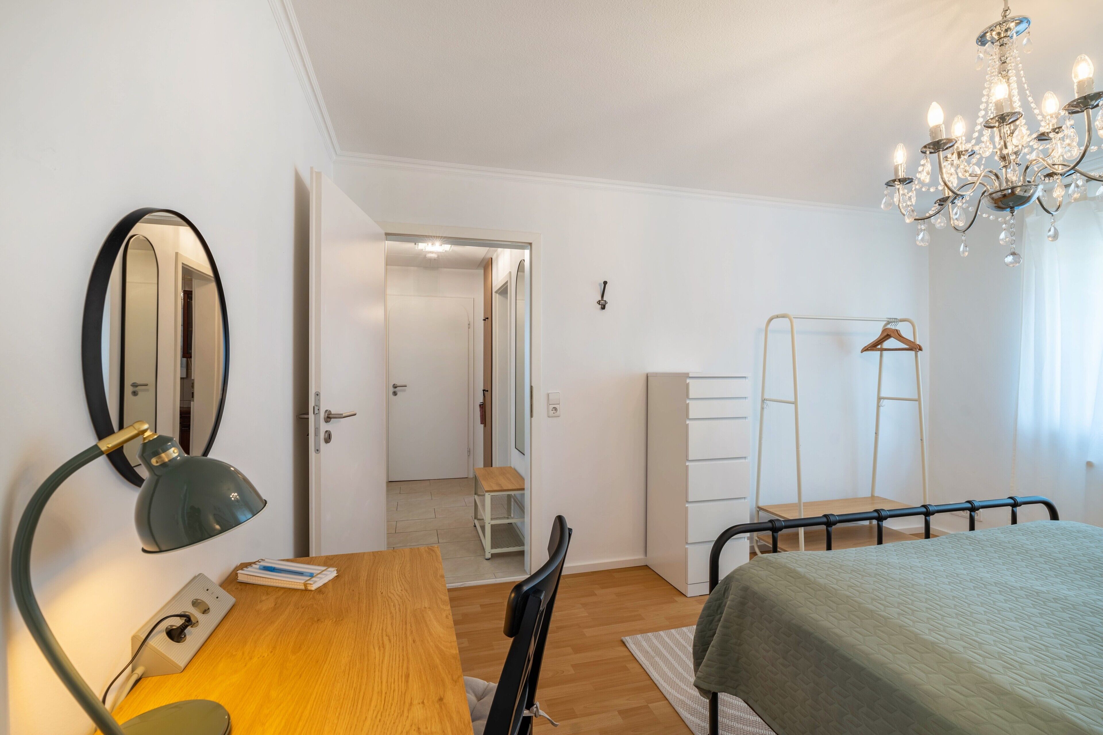 A bedroom with a bed, a desk, a chair, a wardrobe, a mirror, and a chandelier.