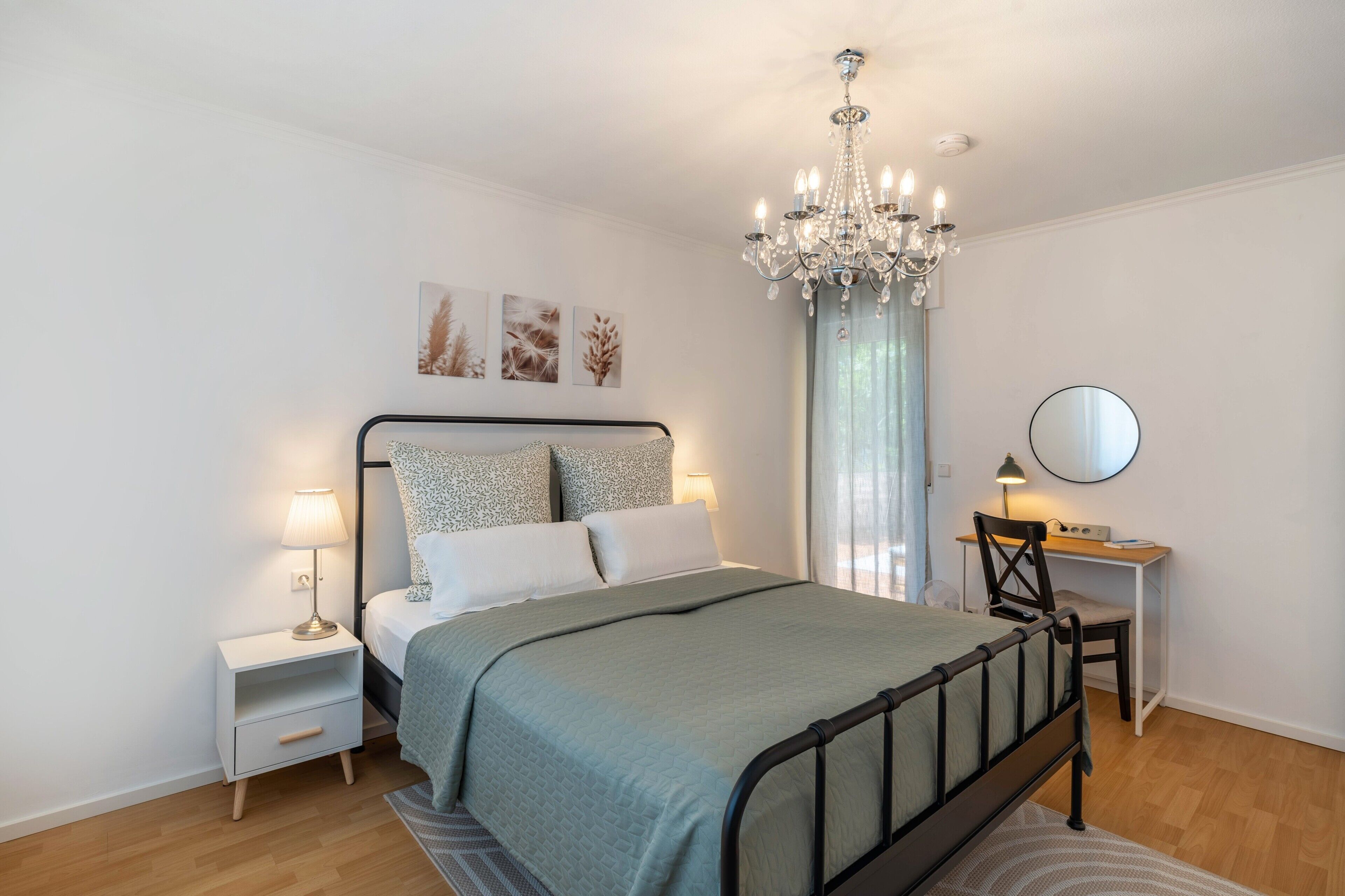 Un dormitorio con cama, mesitas de noche, escritorio, silla, araña y cuadros en la pared.