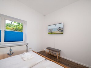 A bedroom with a bed, a window with blue blinds, a radiator, a wall-mounted TV, and a bench. - Modern Ostdeich vacation rental in Büsum with a terrace for the whole family (Büsum)
