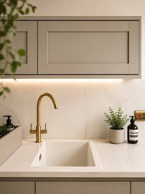 Eine Küche mit einem weißen Spülbecken, einem goldenen Wasserhahn, einer Topfpflanze und einer Flasche Aesop Seife.