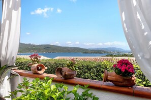 Ein Balkon mit Terrakottatöpfen und Pflanzen mit Blick auf eine Küstenlandschaft mit Hügeln und einem Gewässer.