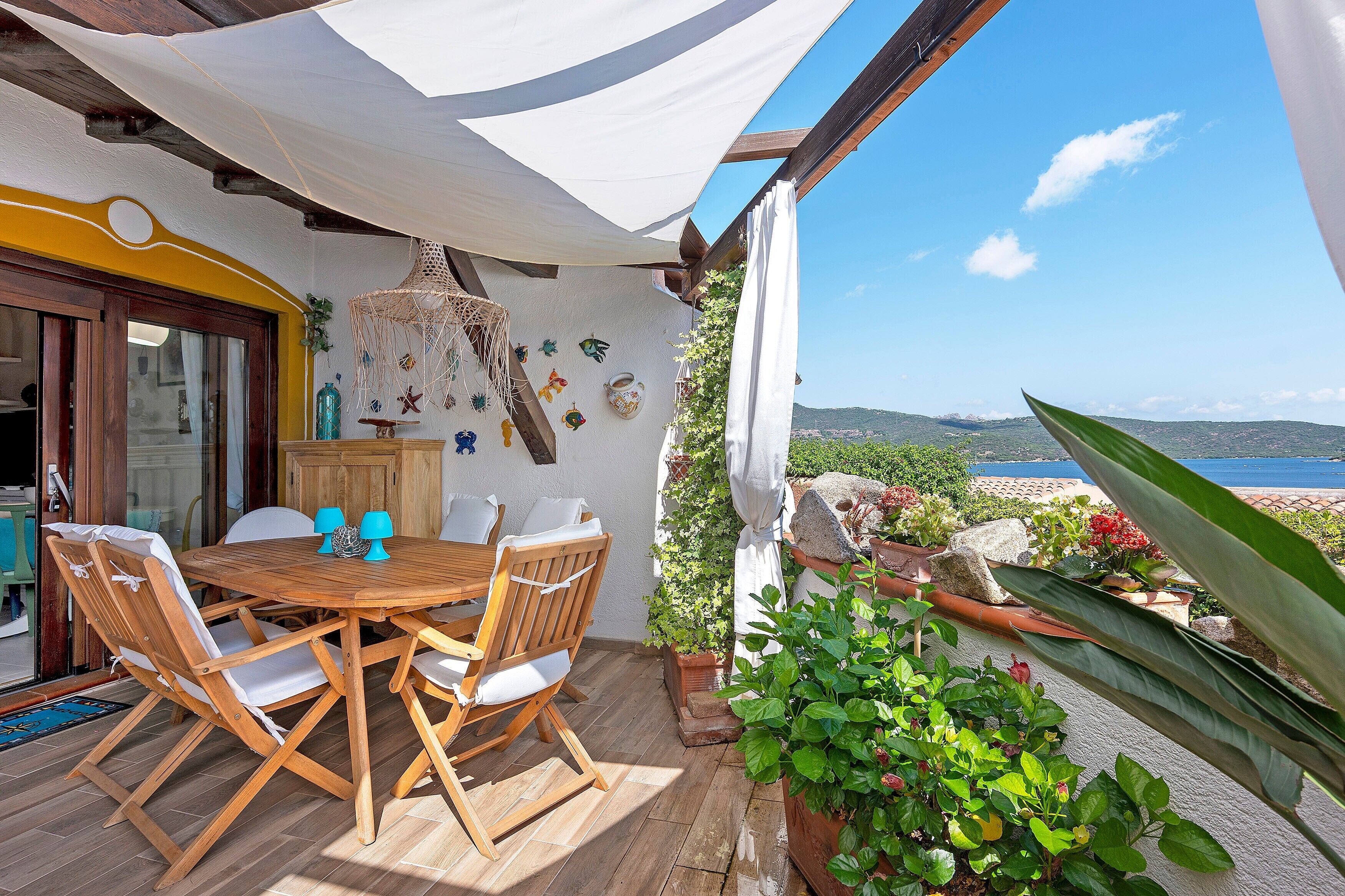 Uma área de refeições ao ar livre com mesa e cadeiras de madeira, um lustre pendurado e vista para o mar.