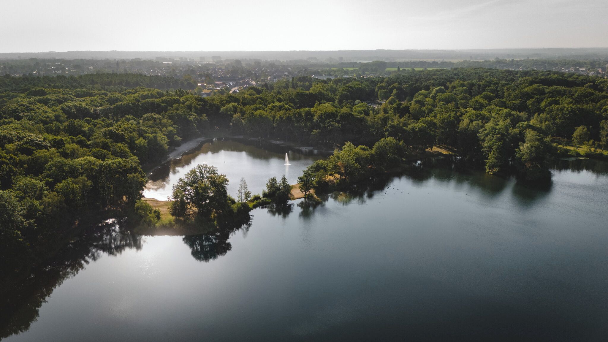 Ein ruhiger See, umgeben von dichtem Grün, mit einer kleinen Insel und einem Springbrunnen.