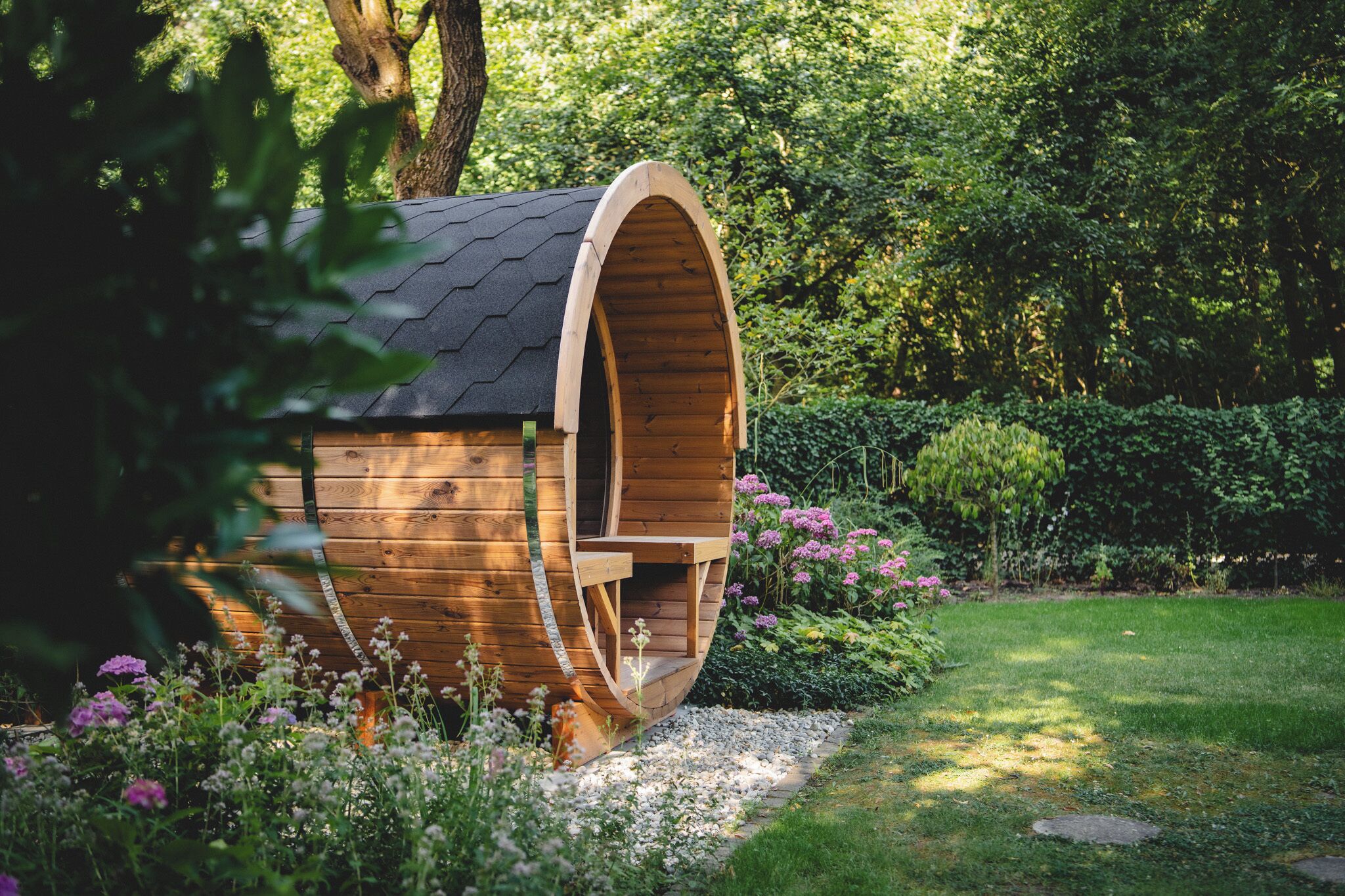 Une structure en bois dotée d’un toit incurvé, entourée d’un jardin où poussent différentes plantes et fleurs.