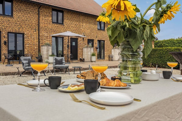 An outdoor dining area with a table set for breakfast, including croissants and orange juice, in front of a brick building with a garden.