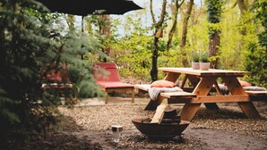 Une table de pique-nique en bois installée à l’extérieur, avec des bancs, entourée d’arbres et bordée d’un chemin de gravier.