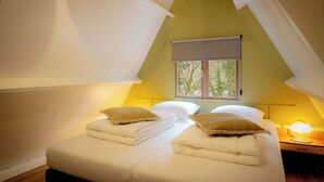 Une chambre avec un plafond en pente, un lit avec une literie blanche et deux oreillers, une fenêtre donnant sur des arbres et une lampe de chevet.