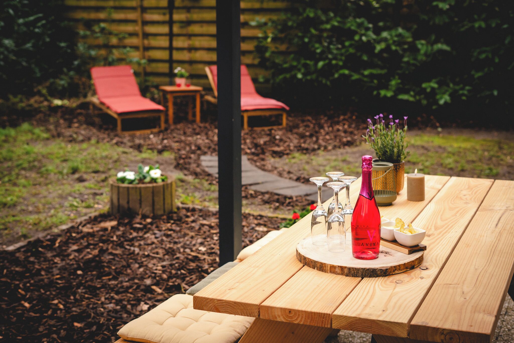 Et træbord med picnic, en flaske og glas, omgivet af en have med et træhegn og røde liggestole.