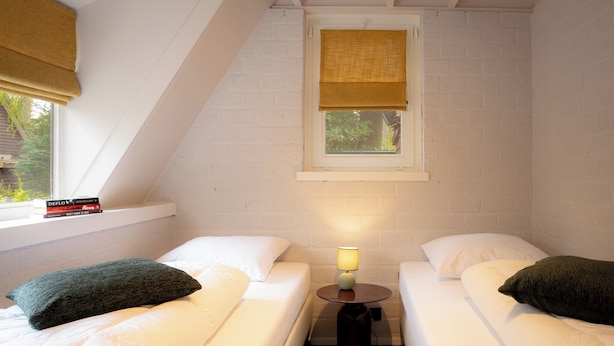Een knus kamertje met twee éénpersoonsbedden, een nachtkastje met lamp en een raam met uitzicht op groen.