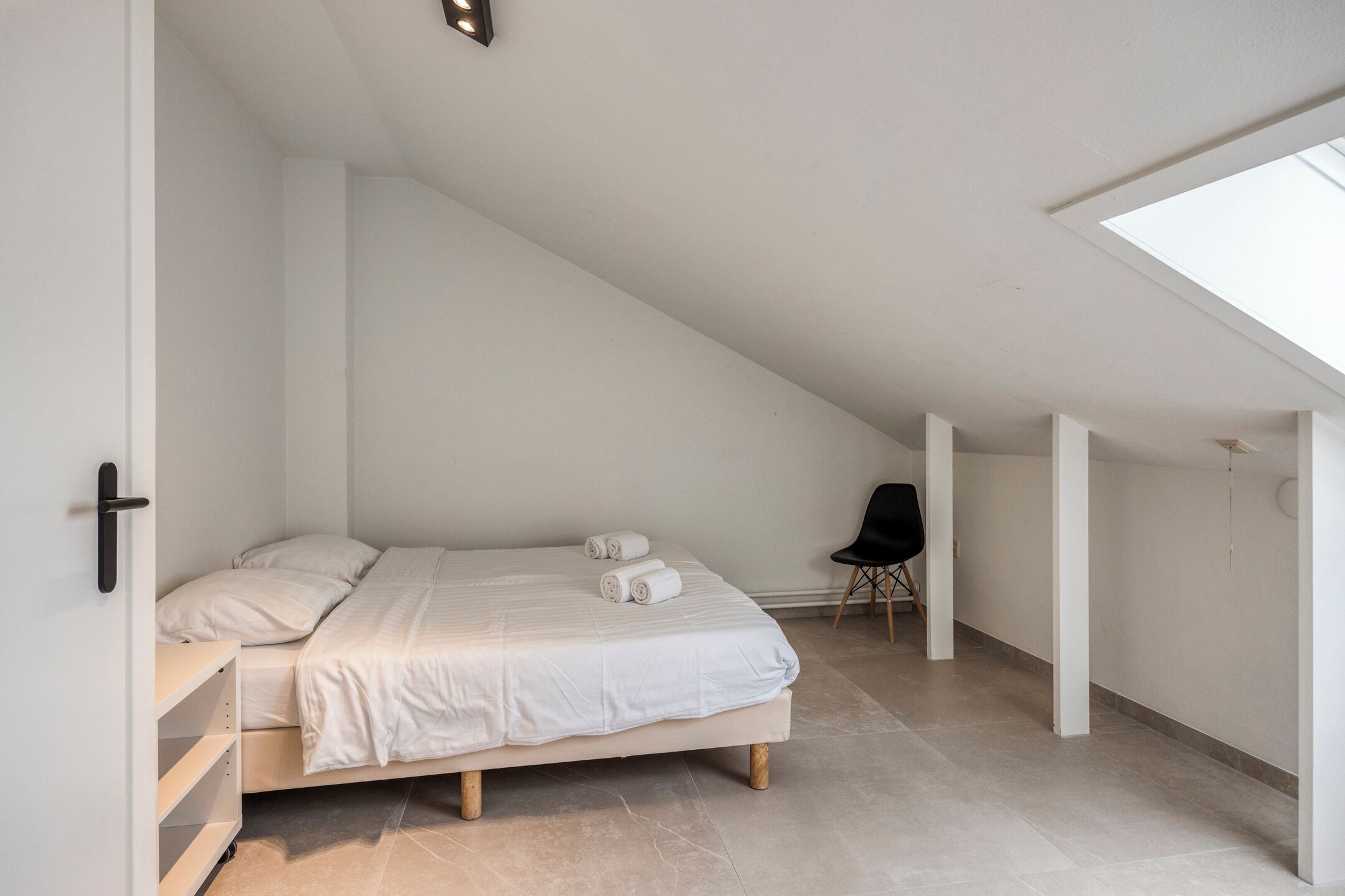 A minimalist bedroom with a sloped ceiling, a bed with white linens, a chair, and a skylight.