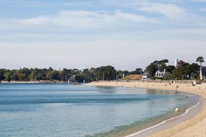 Uma praia com água cristalina, uma faixa de areia e uma fileira de casas ao fundo.