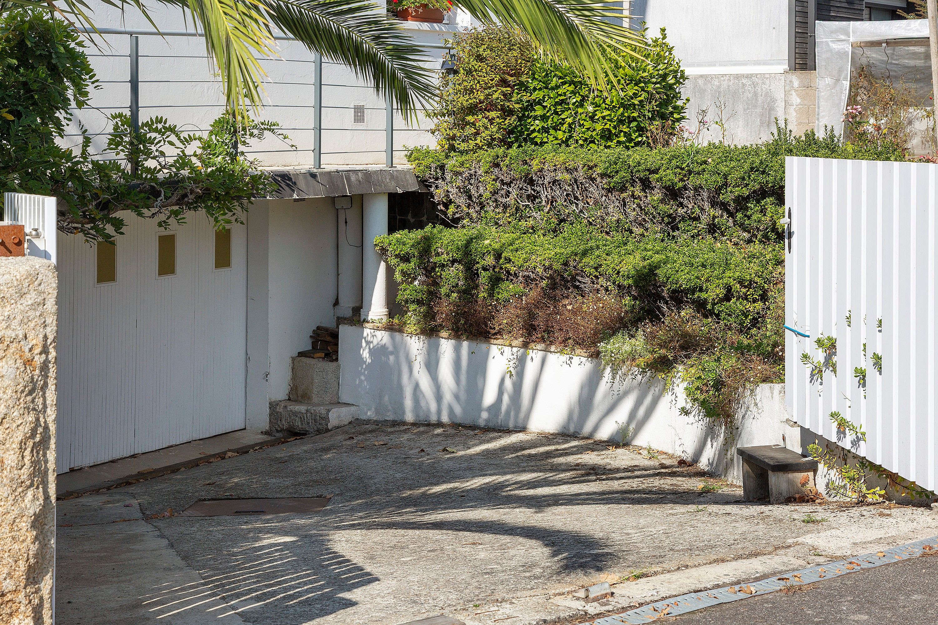 Une ruelle étroite bordée d’une clôture blanche, une petite construction en béton et un feuillage dense.